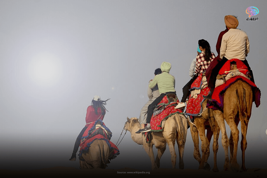 Tourists riding camels across a vast, sandy expanse in the Thar Desert of Rajasthan, with a hazy bright sky.