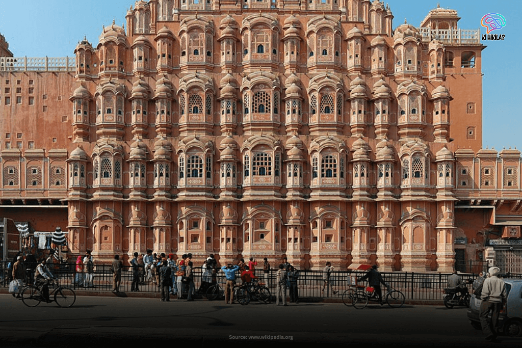 The intricate pink sandstone facade of Hawa Mahal, the Palace of Winds, in Jaipur, Rajasthan, with people and vehicles in front.