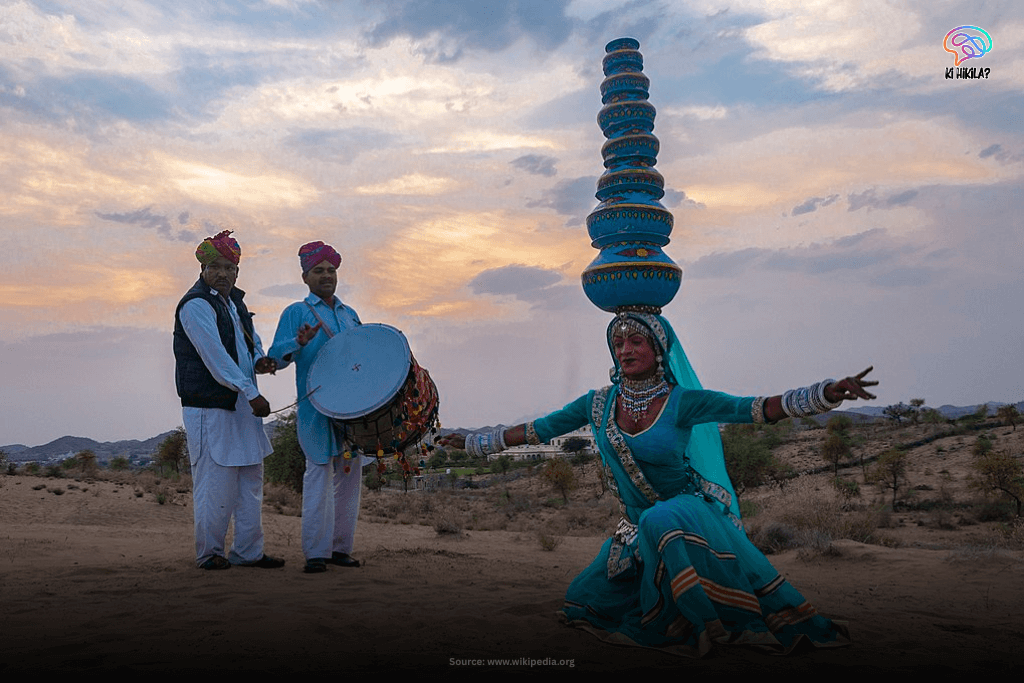 A Kalbelia dancer in a vibrant blue costume with a stack of pots on her head, performing outdoors with two male musicians playing drums in Rajasthan.
