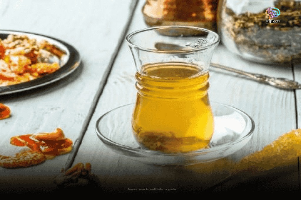 A clear glass filled with golden Kahwa, a traditional Kashmiri tea, sits on a white table with dried fruits and nuts in the background, showcasing the local beverages of Jammu and Kashmir.