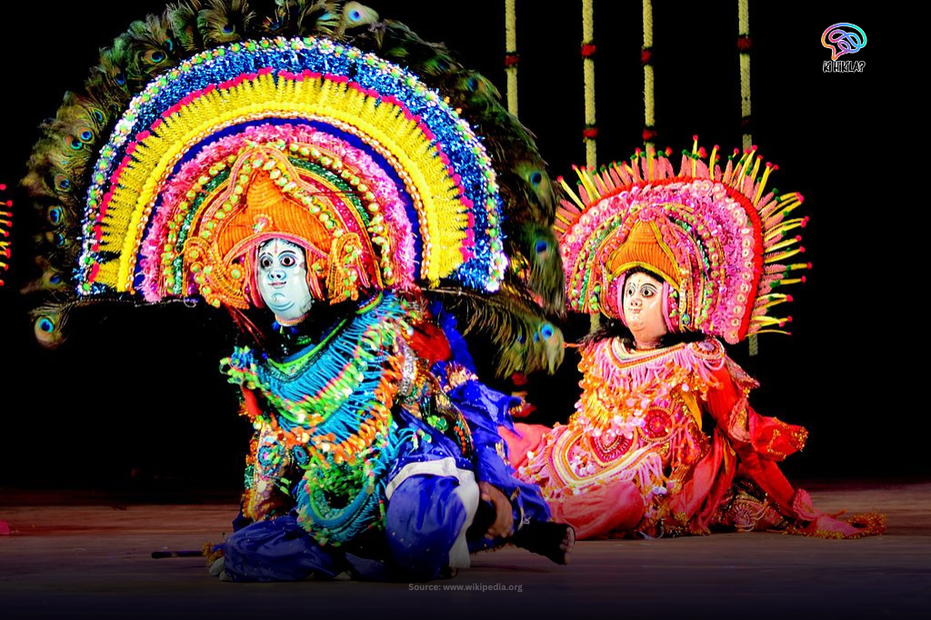 Two Chhau dancers in large, elaborate, and colourful masks and costumes performing on a wooden stage.