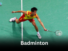 A male badminton player in a yellow and red jersey lunging low on a green court, representing Badminton in Olympic.